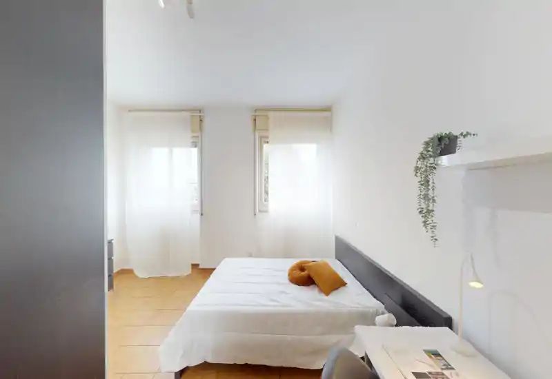 Camera da letto luminosa e minimal con letto matrimoniale, biancheria chiara e luce naturale da due finestre; una scrivania e una mensola rendono l'ambiente funzionale.