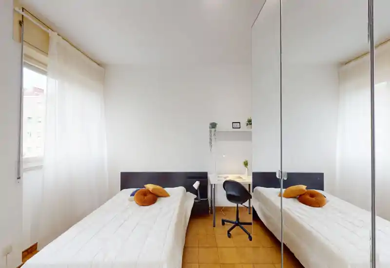 Camera da letto luminosa e minimal con due letti singoli, biancheria bianca, una piccola scrivania e un armadio con ante a specchio che dona profondità. Grandi finestre con tende leggere offrono molta luce naturale e un'atmosfera pulita e rilassante, ideale per ospiti o studenti.