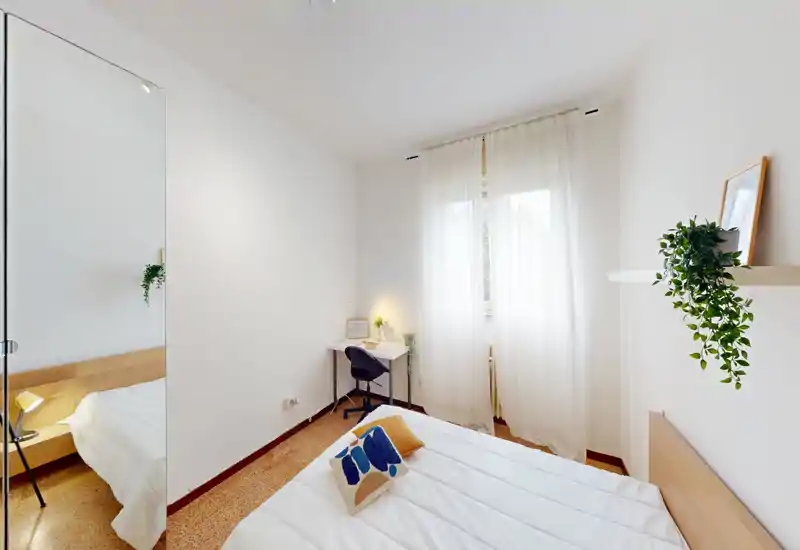 Bright, minimalist bedroom with a neatly made double bed, sheer curtains letting in soft natural light, a small workspace by the window and tasteful plants and cushions for a cozy touch.