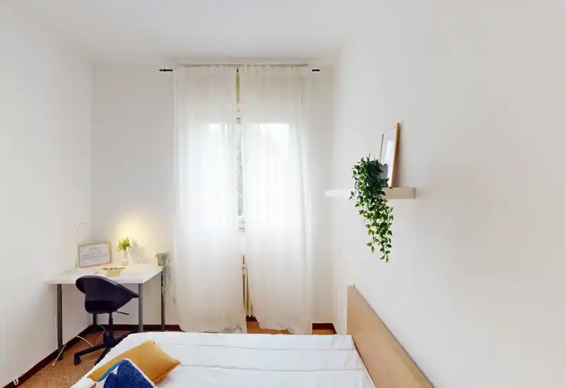 Bright, minimal bedroom with a neatly made bed, sheer window curtains and a small desk in the corner — calm, well-lit space ideal for rest and work.