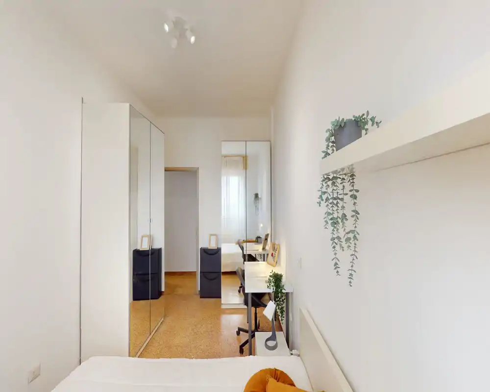 Bright, minimalist bedroom with a white bed, mirrored wardrobe and a small desk. Natural light and simple greenery create a fresh, airy atmosphere ideal for rentals.