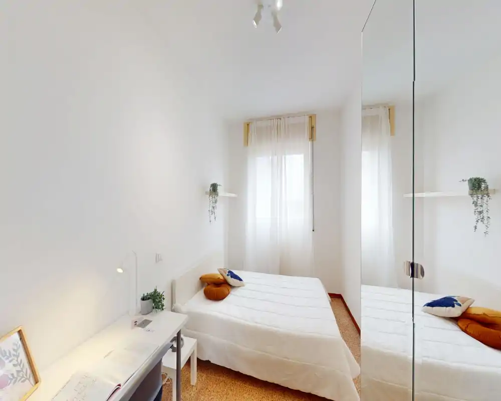 Bright, minimal double bedroom with a neatly made bed, soft decorative cushions and natural light from a sheer-curtained window; includes a desk and mirrored wardrobe that add depth.
