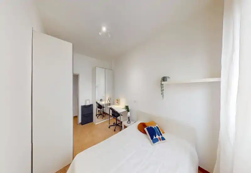 Chambre lumineuse et minimaliste avec un lit bien fait, un bureau et une armoire. Murs blancs, lumière naturelle douce et touches décoratives chaleureuses.