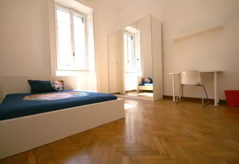 Bright, minimalist bedroom with a low white bed, decorative pillow and mirrored wardrobe; natural light floods the warm parquet floor.
