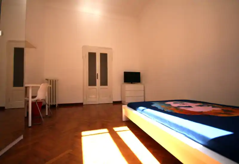 Bright, minimalist bedroom with a low platform bed, wood parquet flooring and sunlight casting across the room. Simple desk and small dresser with TV complete the uncluttered space.
