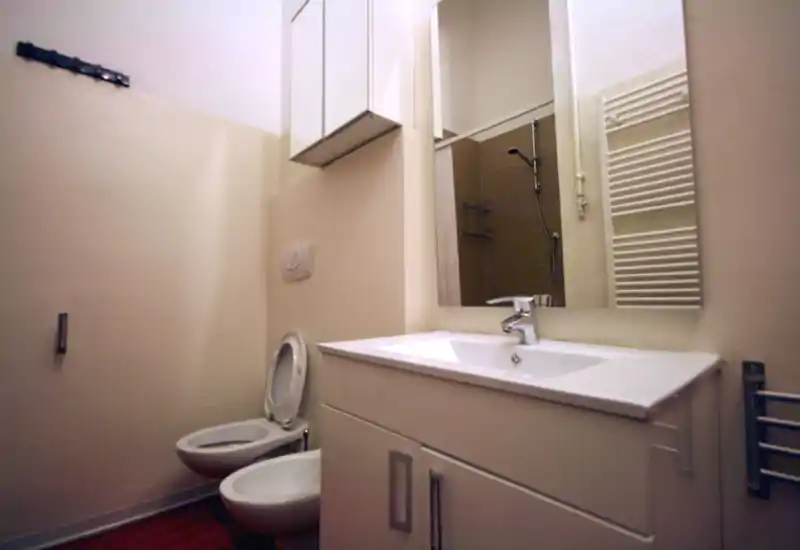 Baño compacto con inodoro suspendido, bidé, mueble con lavabo y espejo que refleja la zona de ducha; tonos crema y sanitarios modernos.