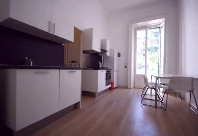 Modern bright kitchen with white cabinets, built-in oven and a small dining table by a large window/door letting in natural light.