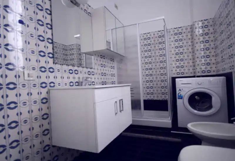 Baño compacto con azulejos blancos y azules con estampado, mueble de lavabo suspendido, mampara de ducha y lavadora de carga frontal. Espacio luminoso y adecuado para un anuncio de alquiler.