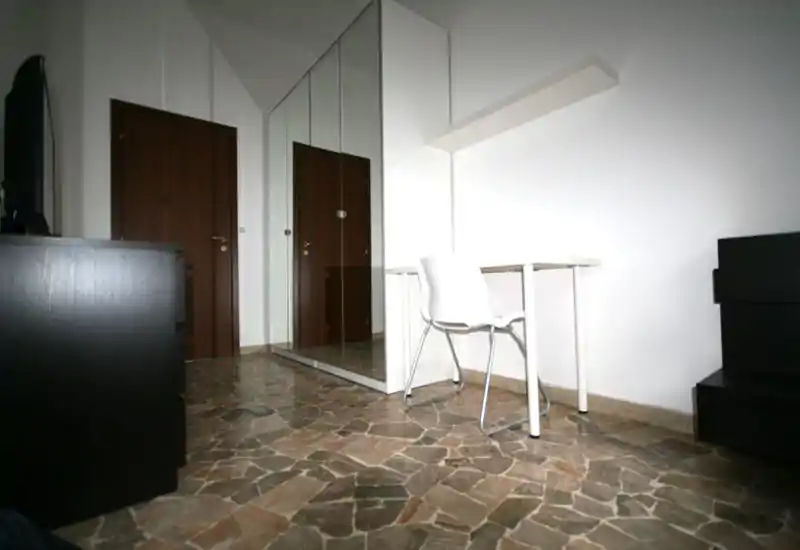 Sparsely furnished living area with a chair and small desk, mirrored wardrobe and stone-patterned floor. Bright white walls and minimal decor create a clean, utilitarian space.