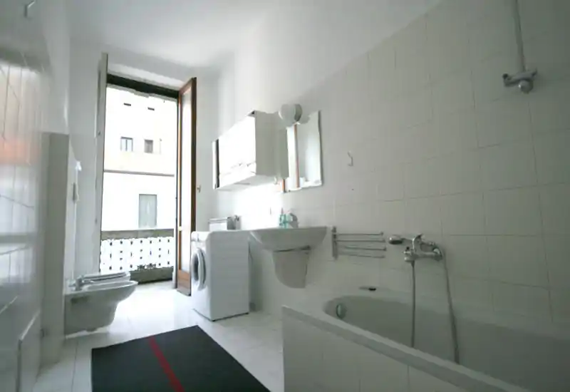 Bright, modern bathroom with a bathtub, wall-mounted sink, bidet, toilet, and a washing machine near a balcony door letting in natural light.