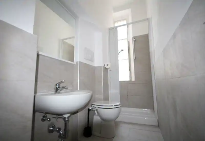 Compact, modern bathroom with a wall-mounted sink, toilet and a tiled shower cubicle beneath a tall frosted window. Neutral light-coloured tiles and clean fixtures create a bright, functional space ideal for a rental listing.