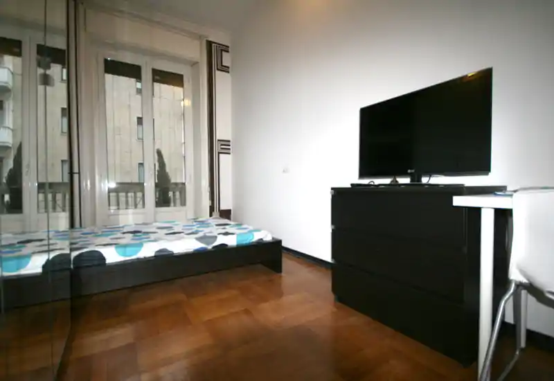 Dormitorio moderno y luminoso con cama doble, amplias ventanas y suelo de parquet. Cómoda negra con televisor y armario con espejo que crean un aspecto limpio y contemporáneo, ideal para alquileres.