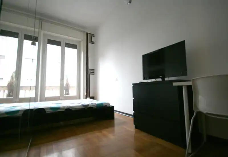 Dormitorio luminoso y minimalista con cama baja junto a grandes ventanales, suelo de parquet y cómoda con TV — espacio para dormir sencillo y aireado.