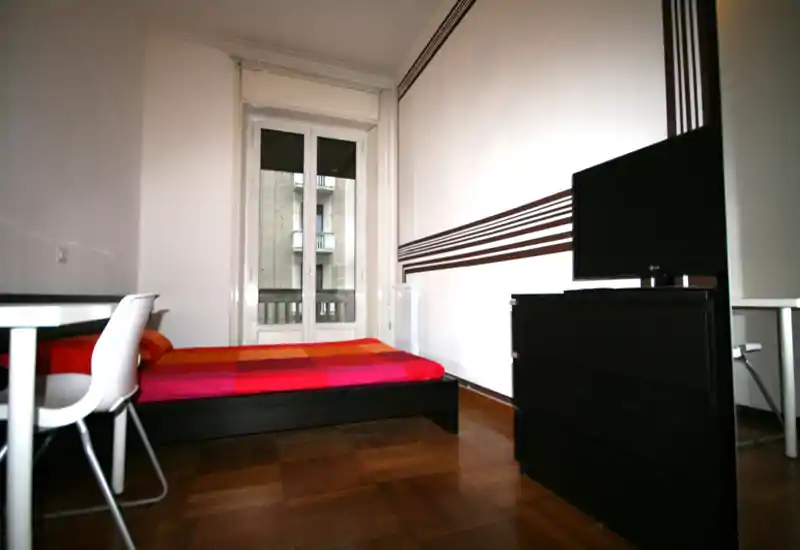 Bright, simply furnished bedroom with a double bed dressed in colorful bedding, desk and chair by the window, and a dresser with TV. Hardwood floors and clean modern lines give a neat, airy feel.