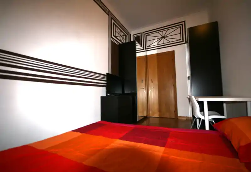 Bright, compact bedroom with a double bed dressed in warm red-orange bedding, decorative wall graphics, a TV on a low cabinet and a small desk — ideal for a cozy city stay.