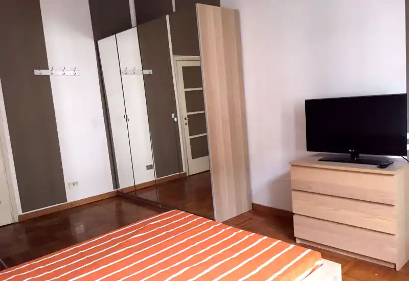 Bright, simply furnished bedroom with a double bed in the foreground, a mirrored wardrobe and a three-drawer wooden dresser topped by a TV.