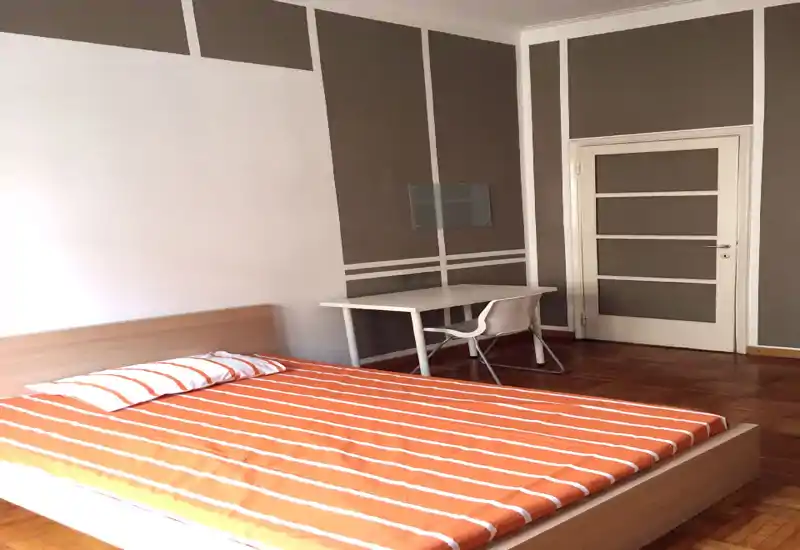 Bright, simply furnished bedroom with a double bed dressed in orange-striped bedding, a compact desk and chair beside the door — a clean and functional space ideal for a single occupant or couple.