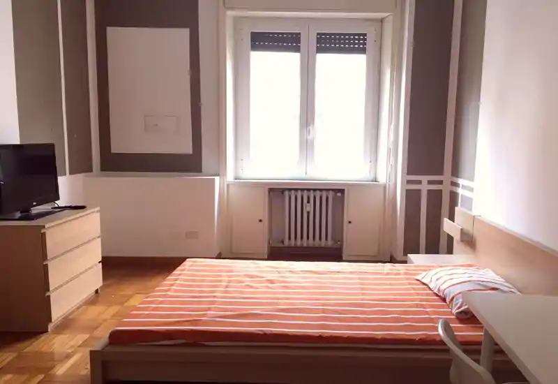 Bright, simply furnished bedroom with a double bed in orange-striped linens, light wood furniture and a large window providing abundant natural light.