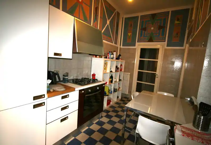 Cocina compacta y luminosa con mobiliario blanco, placa de gas y horno, estantería y mesa de comedor — suelos de baldosa y paneles decorativos en la pared añaden carácter.