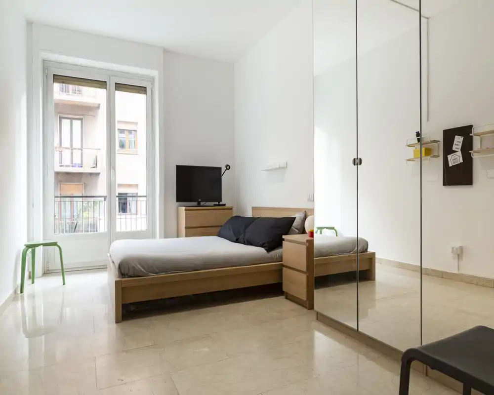 Camera da letto minimal e luminosa con letto matrimoniale in legno, comodini e un grande armadio con ante specchiate. Le ampie finestre garantiscono molta luce naturale e la palette neutra crea un’atmosfera moderna e rilassante.