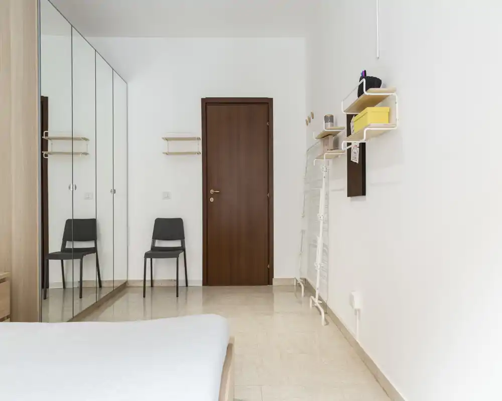 Angolo camera da letto luminoso e minimale con parziale vista del letto, armadio con ante a specchio, una sedia e una porta in legno. Spazio pulito e arioso con toni neutri e mensole a parete pratiche.
