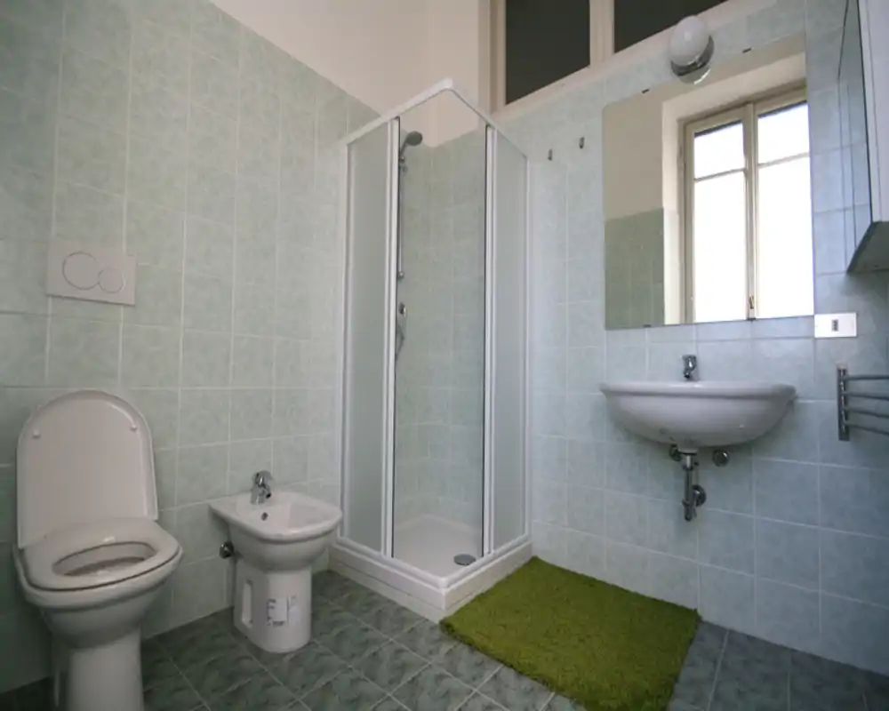 Baño luminoso y funcional con cabina de ducha, inodoro, bidé y lavabo de pared; azulejos verde claro y una ventana aportan luz natural.