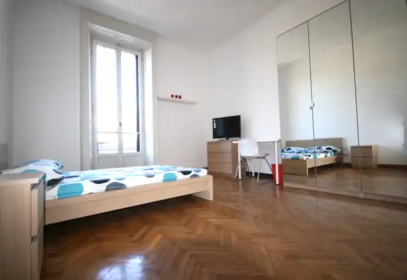 Ampia e luminosa camera da letto con letto basso in legno, biancheria a fantasia e grandi armadi con ante a specchio che riflettono la luce naturale. Pavimento in parquet a spina di pesce e arredo minimal creano un ambiente pulito e arioso, perfetto per gli annunci.