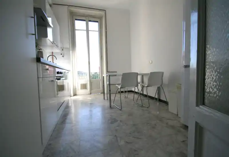 Cucina moderna e luminosa con mobili bianchi, un piccolo tavolo con sedie e una grande porta finestra che fa entrare molta luce naturale.