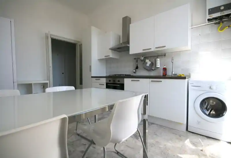 Cucina moderna e luminosa con pensili bianchi, forno a incasso, piano cottura a gas e lavatrice; tavolo con piano in vetro e sedie bianche in primo piano.