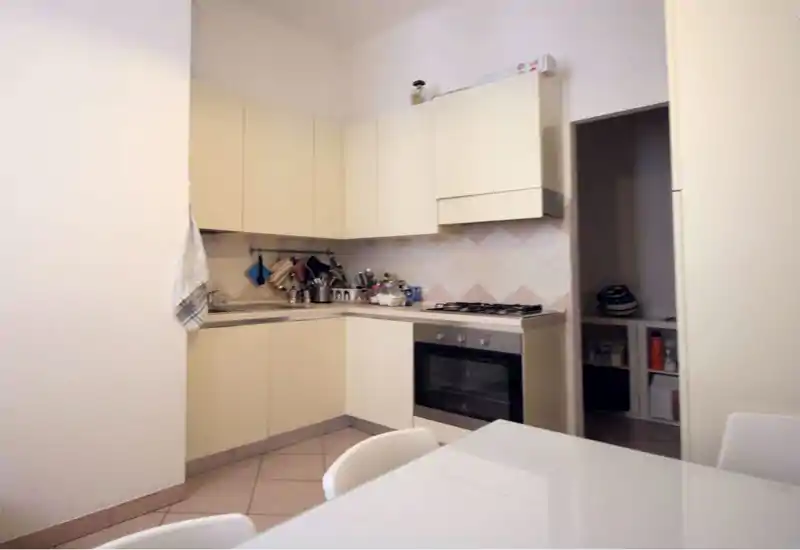Cucina angolare compatta e luminosa con pensili crema, forno incassato e piano cottura a gas. Spazio ordinato e funzionale con utensili e asciugamano visibili, perfetto per annunci di appartamento.