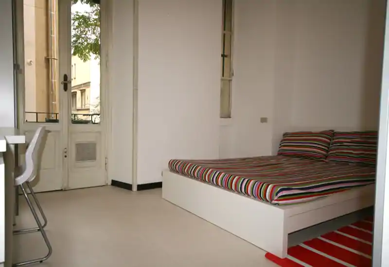 Chambre moderne et lumineuse avec un lit bas blanc à plateforme, linge de lit rayé coloré, tapis rayé rouge et lumière naturelle provenant d'une porte-fenêtre.