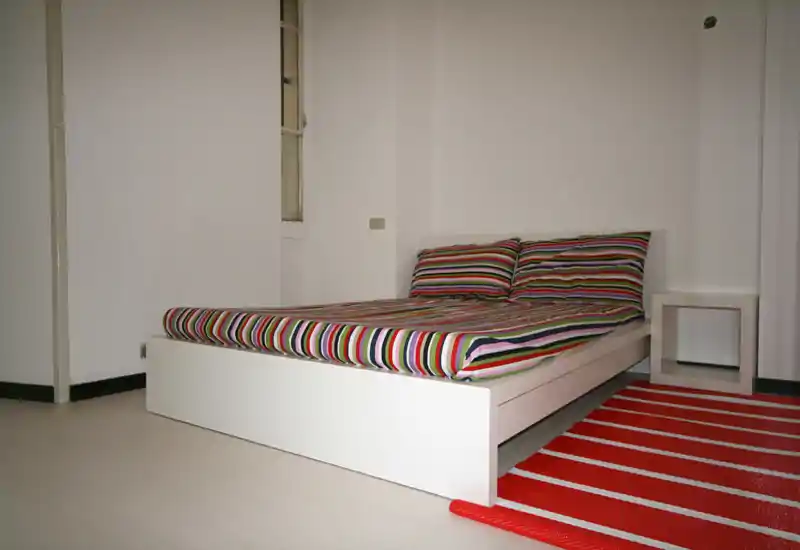 Chambre lumineuse et minimaliste avec un lit bas blanc sur plateforme, linge de lit à rayures colorées et tapis rouge rayé ; table de chevet simple et murs blancs pour une ambiance contemporaine et apaisante.