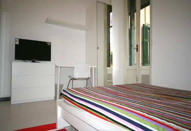 Chambre moderne et lumineuse avec lit à rayures au premier plan, commode blanche avec télévision, bureau et chaise, et grandes fenêtres apportant la lumière naturelle.