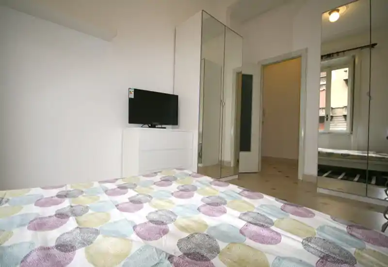 Dormitorio moderno y luminoso con colcha estampada, armarios con espejo y una cómoda pequeña con televisor. La habitación es limpia y aireada con luz natural por la ventana.