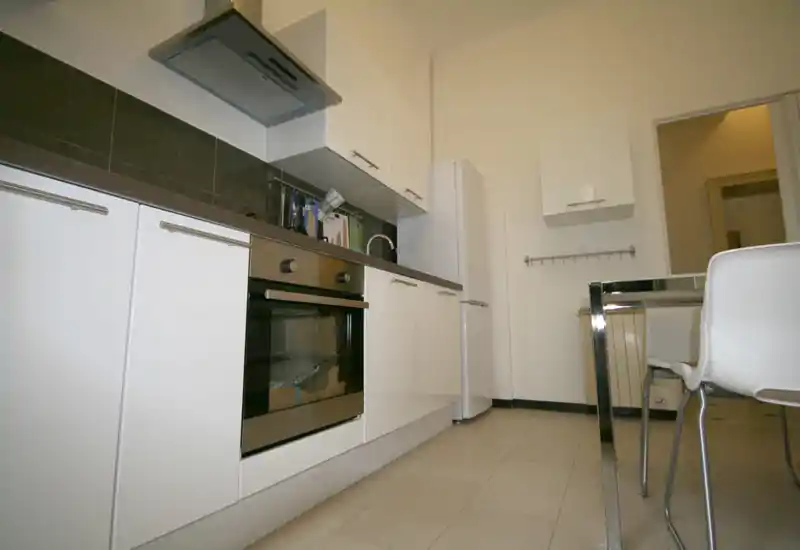 Cocina moderna y compacta con muebles blancos, horno empotrado, frigorífico y una pequeña mesa — espacio luminoso y limpio, ideal para un piso en la ciudad.