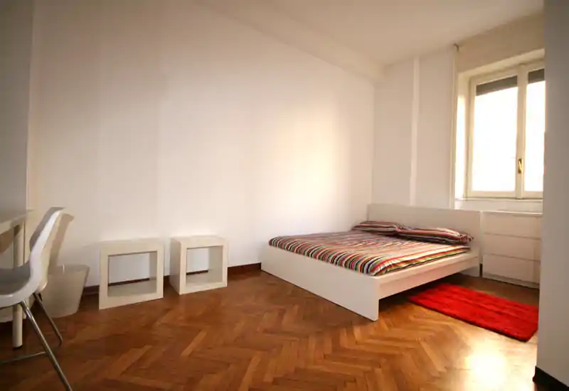 Dormitorio luminoso y minimalista con cama baja blanca, ropa de cama a rayas y una alfombra roja junto a la ventana. Suelo de parquet en espiga y mobiliario sencillo aportan un ambiente limpio y aireado, ideal para anuncios.