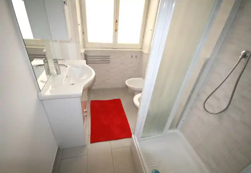 Baño compacto y bien iluminado con plato de ducha, lavabo con mueble, bidé y una ventana luminosa. Azulejos neutros y una alfombrilla roja que aportan un aspecto limpio y moderno.