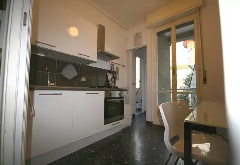 Cocina compacta y moderna con armarios blancos, horno y placa integrados, mesa pequeña con sillas y acceso a un balcón que aporta luz natural.