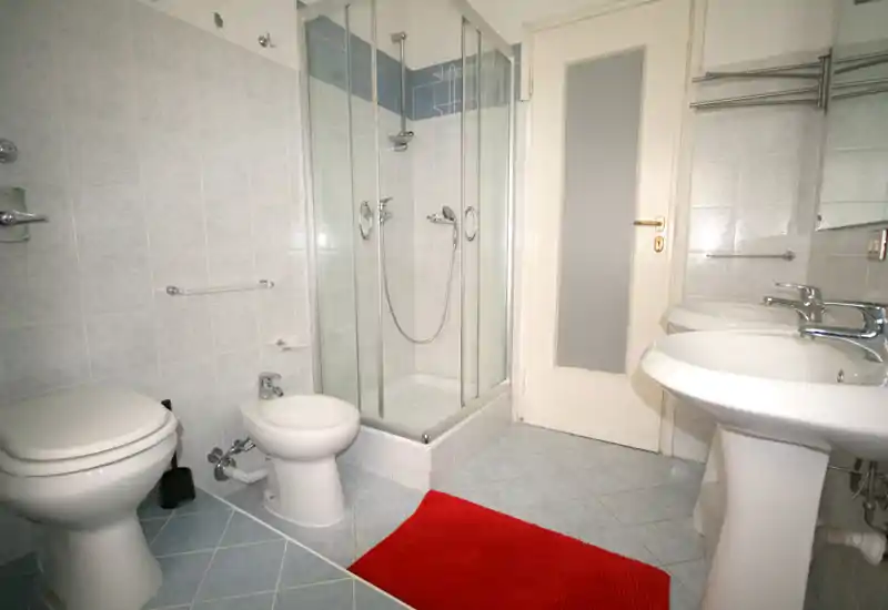 Bagno luminoso e moderno con doccia angolare in vetro, bidet, WC e doppio lavabo; piastrelle azzurre chiare e un tappetino rosso per contrasto.