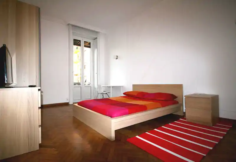Ampia camera da letto luminosa con letto moderno in legno e biancheria dai toni rosso-arancio. Grande porta finestra che fornisce luce naturale e un piccolo angolo scrivania vicino al balcone.