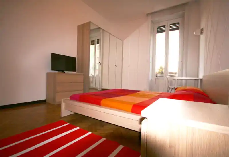 Camera da letto luminosa e moderna con letto basso in legno coperto da biancheria rossa e arancione, pavimento in parquet, armadio con specchi e comò con TV: ambiente pulito e accogliente.