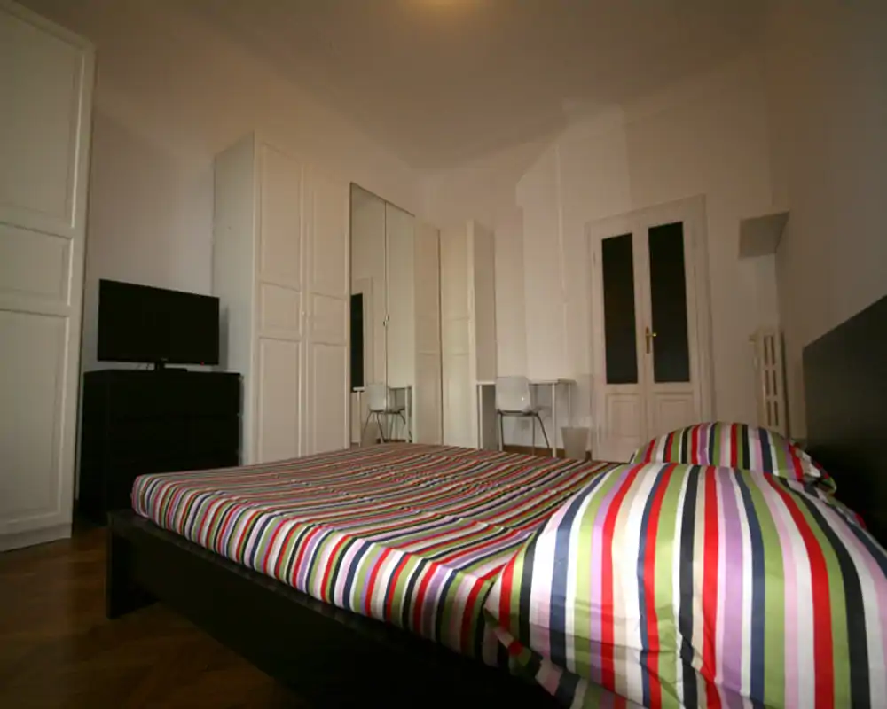 Ampia camera da letto con letto basso e biancheria a righe colorate, una parete di armadi bianchi con specchi, una piccola scrivania e TV — luminosa, moderna e dal design essenziale.