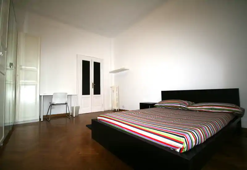 Ampia camera da letto minimalista con letto basso nero su pedana, biancheria a righe colorate, pavimento in parquet e pareti bianche che creano un ambiente luminoso.
