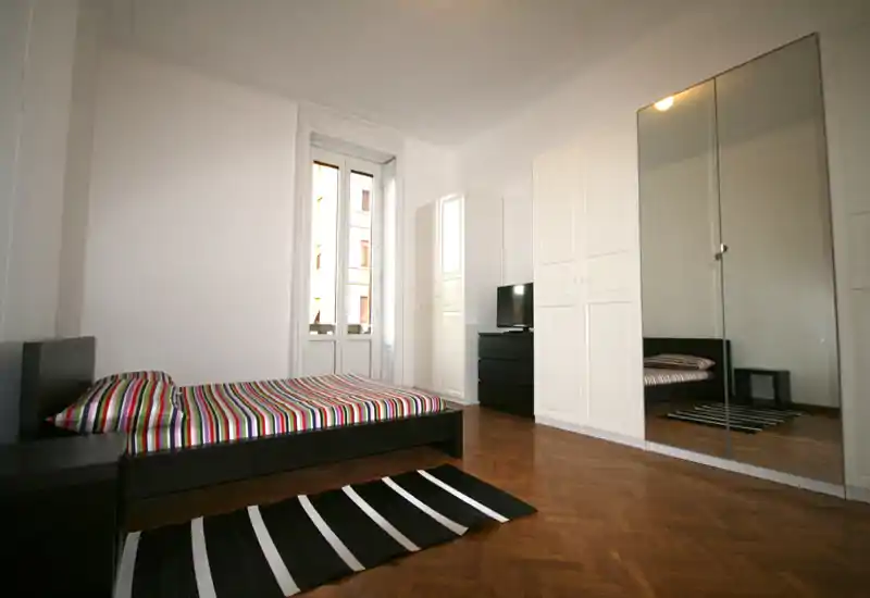 Camera da letto spaziosa e luminosa con letto basso, copriletto a righe e grande guardaroba con specchi che amplifica la percezione dello spazio.