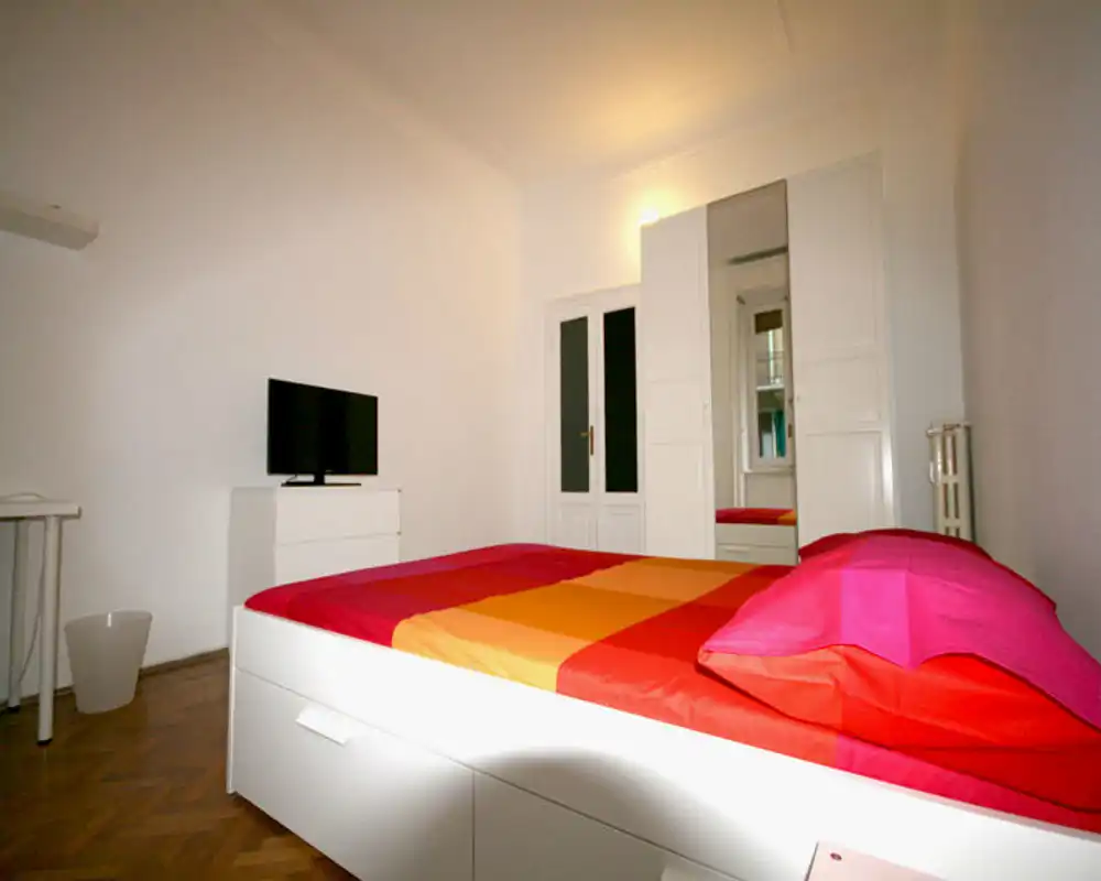 Dormitorio luminoso y minimalista con una amplia cama blanca y ropa de cama naranja y rosa, un armario con espejo y una pequeña TV — limpio, moderno y bien iluminado.