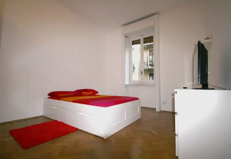 Dormitorio luminoso y minimalista con cama plataforma blanca, ropa de cama colorida y ventana que aporta luz natural. Suelo de parquet y alfombra roja completan el aspecto moderno y sencillo.