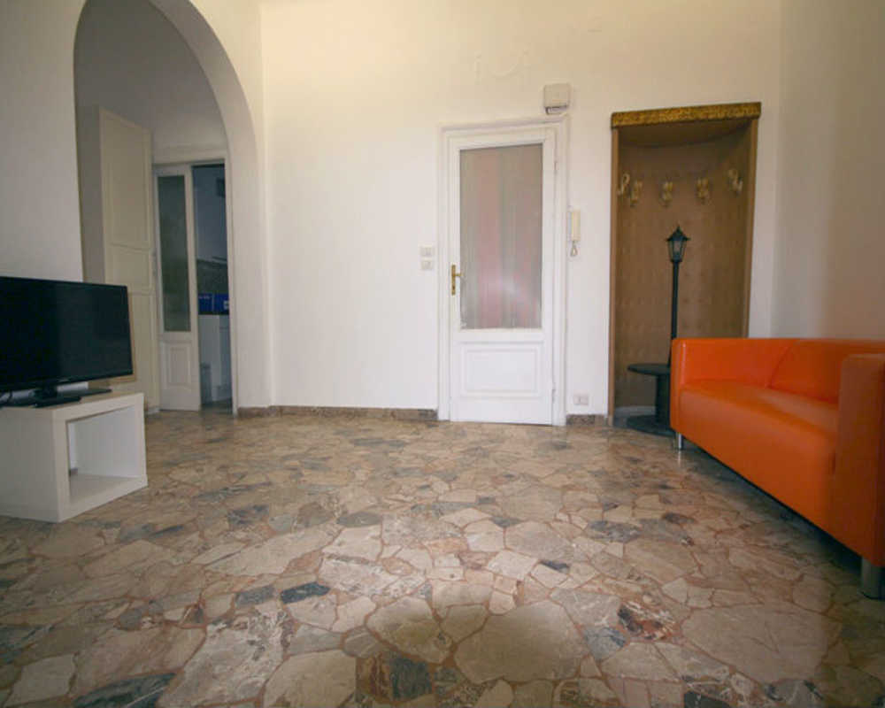 Ampio soggiorno con pavimento in pietra chiara, divano arancione e angolo TV; layout semplice e arioso con arco d'ingresso e nicchia appendiabiti.