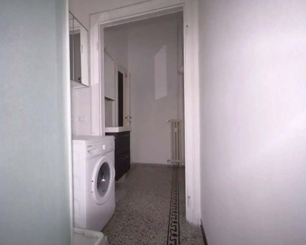 Corridoio d'ingresso stretto con lavatrice visibile e porta che conduce a una stanza interna; pavimento piastrellato con bordo decorativo e pareti bianche neutre.