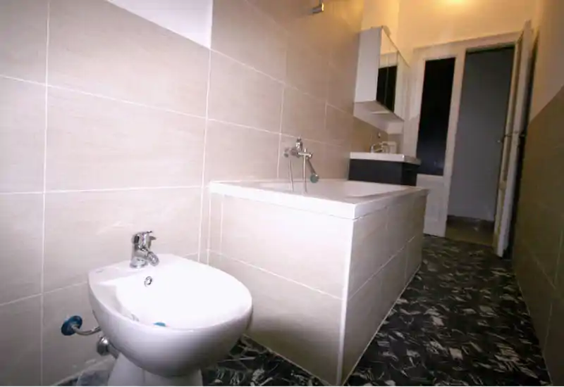 Baño moderno alicatado con bañera, bidé y mueble de lavabo mural; suelo oscuro con dibujo y azulejos neutros en paredes para un aspecto limpio y contemporáneo.