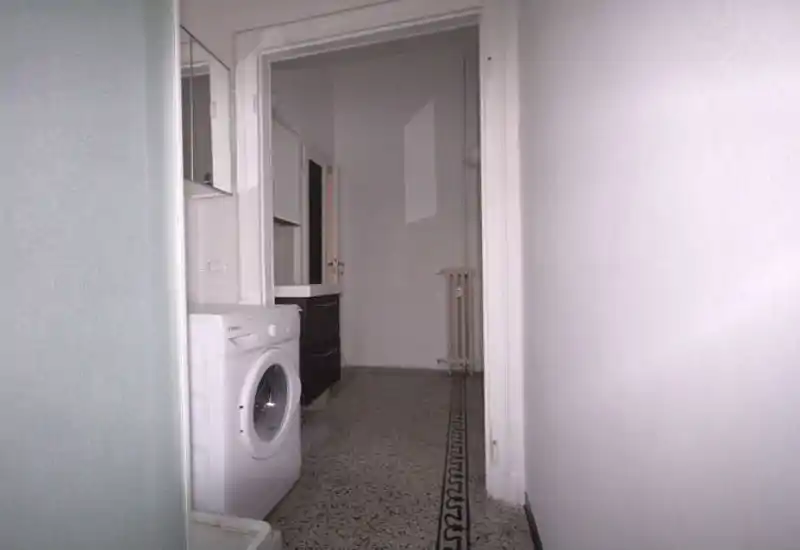 Pasillo de entrada estrecho con lavadora visible y puerta hacia una habitación interior; suelo con cenefa decorativa y paredes blancas neutras.
